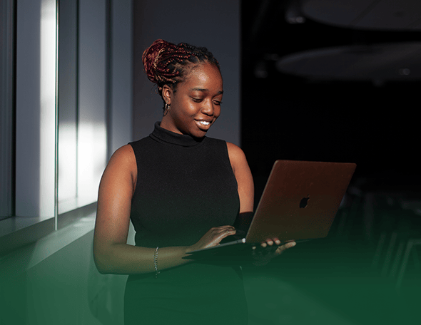 A smiling person in a sleeveless black top stands by a window, looking at a laptop. The daylight streams in, partially illuminating them against a dark background. Their hair is styled in braids and pulled back. A green gradient overlays the bottom of the image, subtly hinting at their School of Business affiliation.