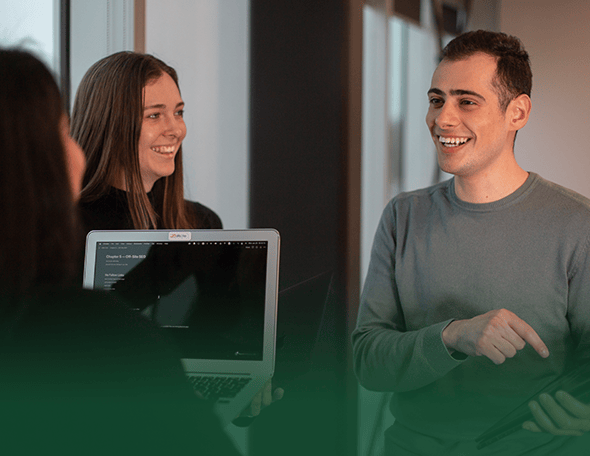 Three individuals are engaged in a lively conversation in an office setting from the school of business. One person is holding a laptop, another is gesturing with their hand, and the third person is facing them. They are all smiling warmly, creating an atmosphere of collaboration and camaraderie.