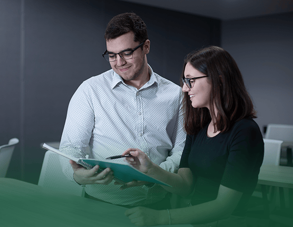 Two people from the school of business are in a discussion. One is holding and looking at a notebook while the other is pointing at it with a pen. Both are wearing glasses and smiling. The background features a dimly lit room with white chairs. The bottom part of the image is gradient green.