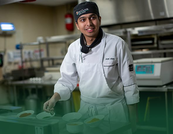 A chef in a white uniform and black hat stands in a commercial kitchen. He is holding a spoon over bowls filled with different ingredients and smiling. The background, featuring kitchen equipment and a fire extinguisher on the wall, also displays a banner for St. Clair Media Arts.