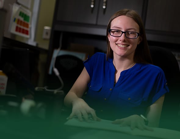 A woman with long hair and glasses, wearing a blue shirt, sits at a desk smiling. She is working at a computer in the School of Business, with office supplies and cabinets visible in the background.