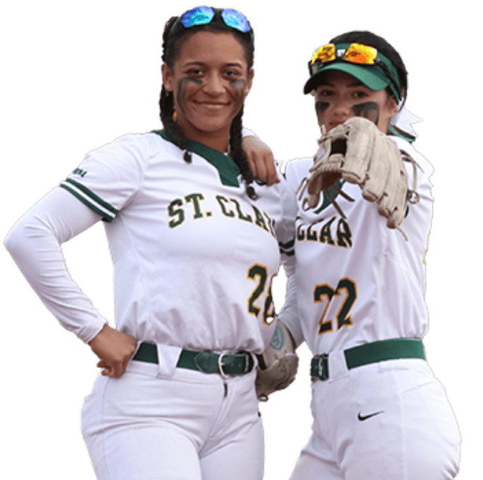 Two female softball players in white uniforms with "St. Clair" and green trim stand together. One player has her arm around the other, who is pointing forward with a glove on her hand. Both have black face stripes, conveying a confident and competitive spirit perfect for recruitment in Sept 2024.