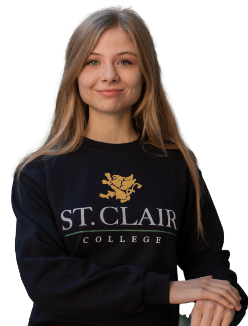 A young woman with long blonde hair is standing and smiling at the camera, embodying the spirit of recruitment for Sept 2024. She is wearing a dark sweatshirt with a yellow gryphon and the text "ST. CLAIR COLLEGE" on it, one arm resting on the other. The background is transparent, perfect for a new slider.